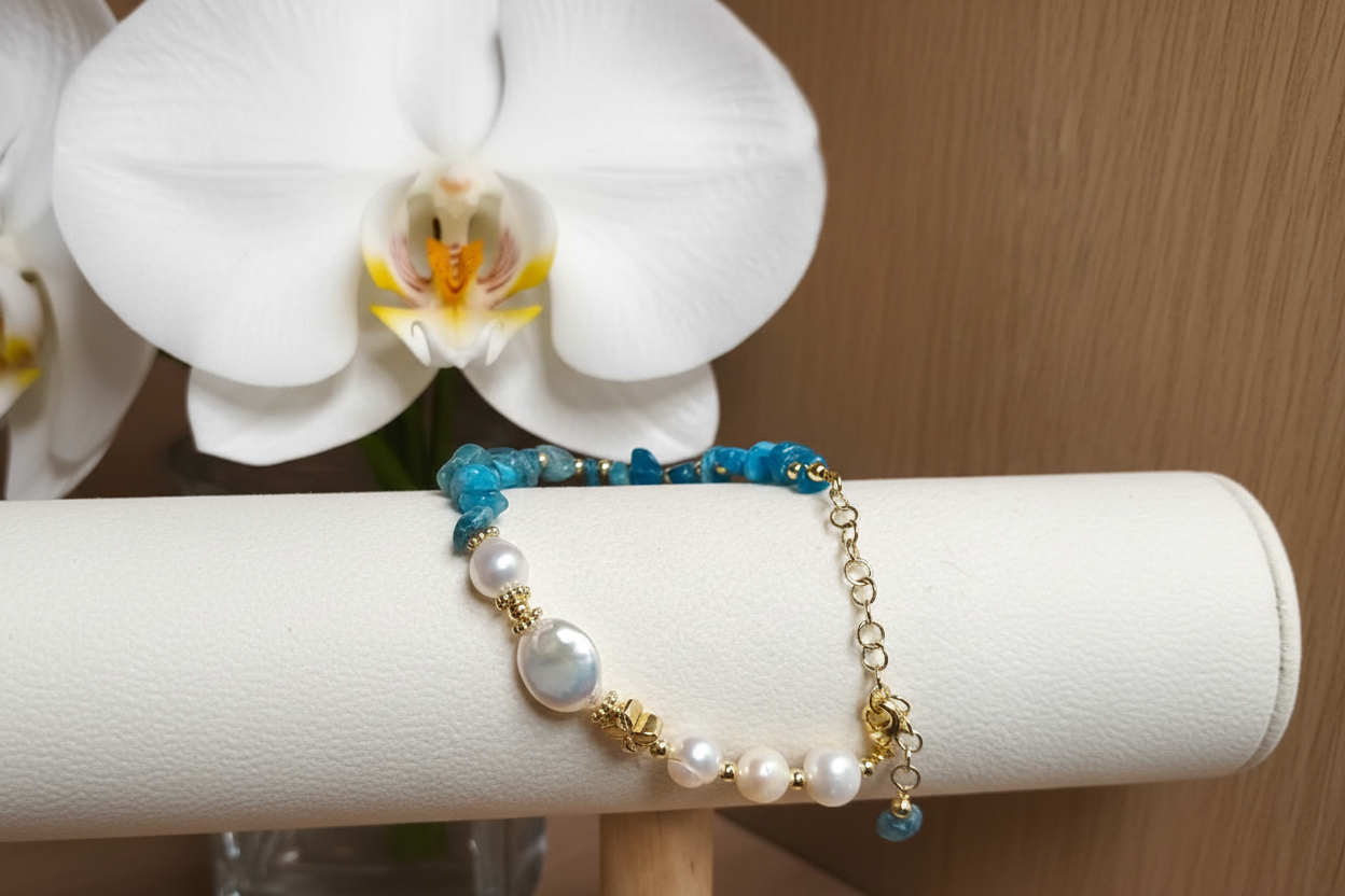 Bracelet with blue beads, pearls, and gold accents on a white surface with flowers in the background